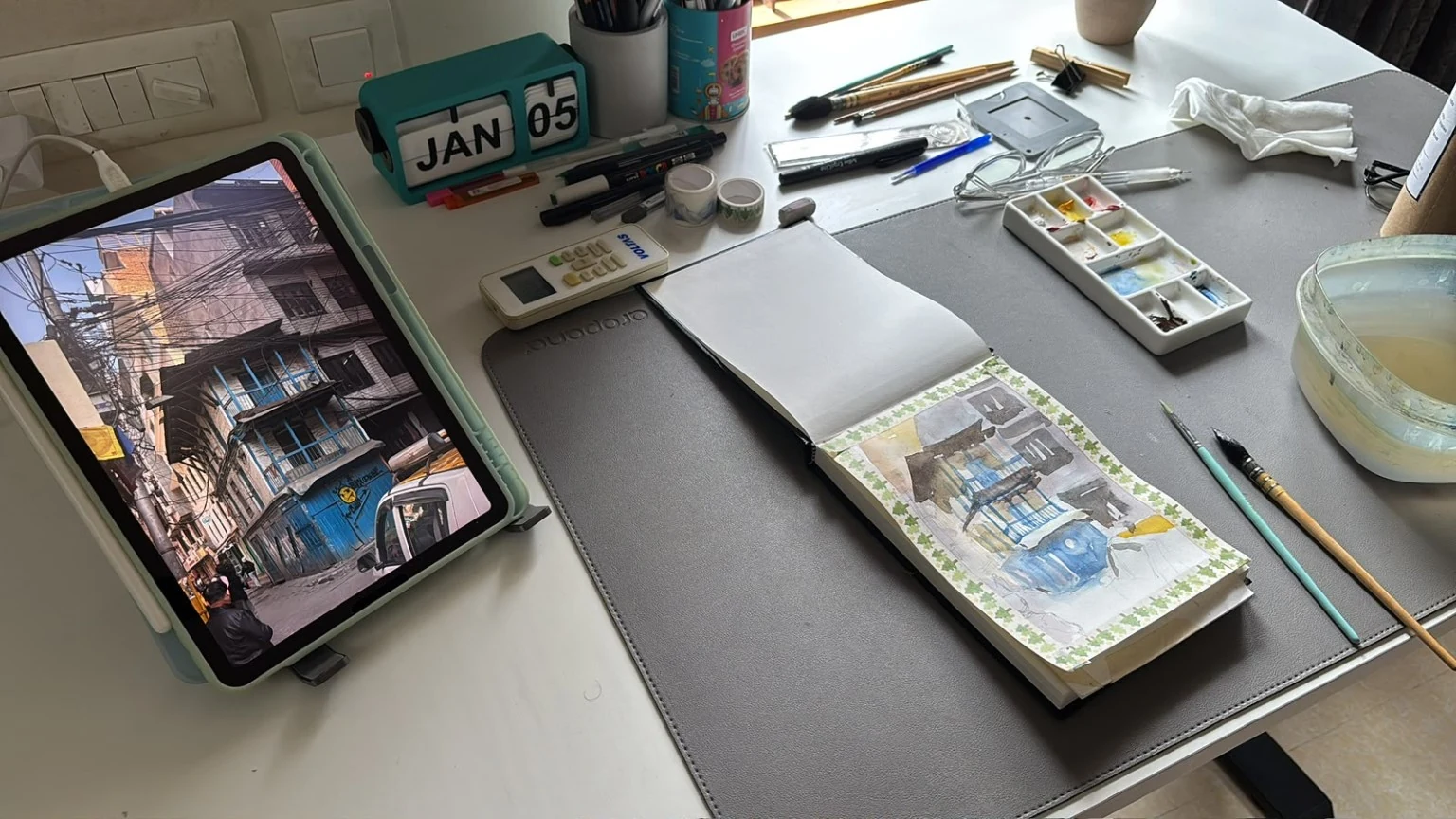 Working table with an open sketchbook, drawing tools, paint palette and tablet